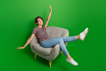 Full size portrait of overjoyed gorgeous girl sit comfy chair raise hands rejoice isolated on green...