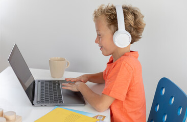 Little boy with white headphones and blonde hair, plays and has fun with a laptop