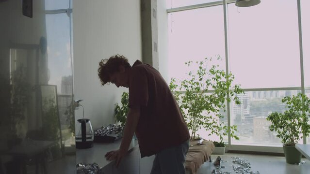 Hungover gen Z man removing tinsel from kitchen countertop, leaning on it and drinking water from bottle while recovering after party at home