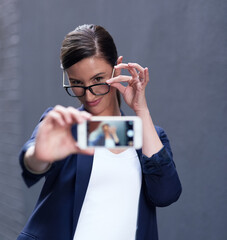 Businesswoman, selfie and cellphone by dark background for online company, social media and pride. Confident female person, technology and glasses with emoji face for internet and profile picture