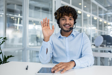 Hindu male wearing headset with microphone and raising hand in greeting gesture while sitting by...
