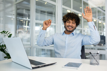 Curly islamic man in blue shirt raising arms for stretching back while sitting at workspace with...