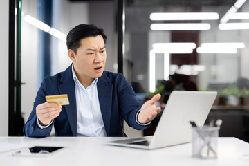 Confused asian male in shirt and jacket looking at computer screen and holding bank card in hand....