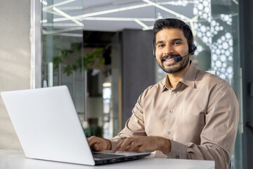 Satisfied muslim male in headset with micro using contemporary gadget by white desktop at workspace. Smiling bearded guy in beige shirt typing on keyboard of pc and smiling widely at camera.