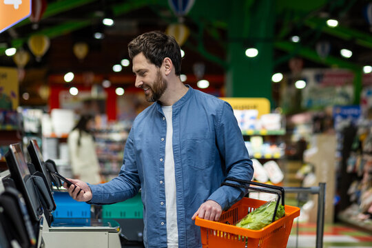 Satisfied customer of food store tapping modern gadget to electronic device while carrying orange basket. Relaxed man in denim shirt enjoying benefits of using banking application for remote payment.