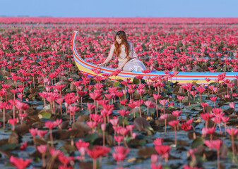 A young woman in Thai national costume sits on a boat touching a lotus flower. It is in a pink...
