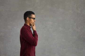 Pensive african american customer thinking over special offer, touching chin looking forward on grey studio copy space. Handsome young man in casual shirt and glasses side view portrait
