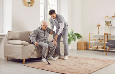 Portrait of a young male nurse helping a senior patient to get off the couch with crutches at home. Physiotherapist helping old man in rehab. Health care worker helping an elderly man
