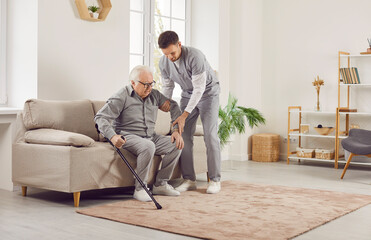 Portrait of a young male nurse helping a senior patient to get off the couch with crutches at home....