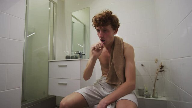 Young hungover man sitting on toilet in bathroom, brushing teeth and wiping mouth with towel while feeling sick next morning after party