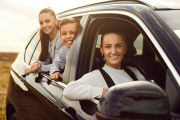 Happy family traveling by automobile. Single mother and her children enjoying road trip together. Beautiful woman driver and kids sitting inside good modern black car, looking out window and smiling