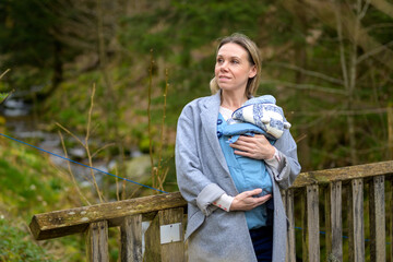 Happy woman looking to side while holding and carrying it in a baby carrier standing on a bridge