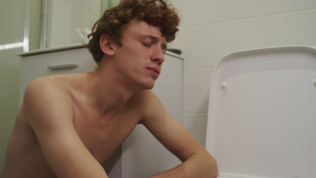 Young sick man feeling nausea leaning on toilet and then resting in bathroom while suffering from hangover in morning at home