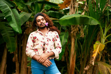Confident bipoc gay man with flower in mouth stands in tropical garden. Fashionable male enjoys...
