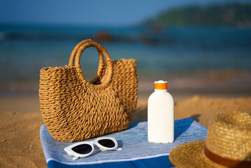 Brown straw bag, white sunscreen bottle, sun hat, white sunglasses on blue towel by sea on sunny...
