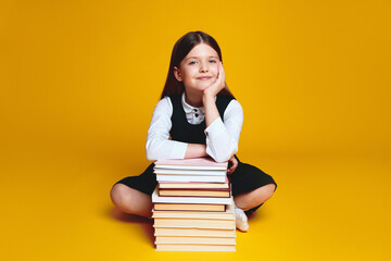 Cheerful elementary schoolgirl in uniform, sitting near bunch of books with crossed legs and hand under chin, isolated on yellow background