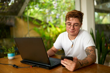 Transgender office worker with tattoos checks smartphone, distracted from laptop. Casual business...