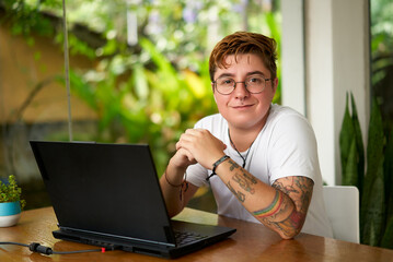 Transgender man with glasses and tattoos works on laptop in bright coworking space. Smiling,...