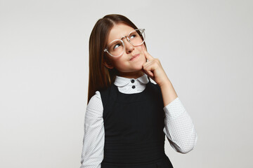 Puzzled little girl wearing school uniform and eyeglasses biting lip and looking away while...