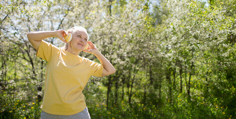  lifestyle portrait senior woman  with gray hair in headphones relaxes in a flowering garden in spring. spends time in nature. Audio healing. meditation. banner. copy space. greyhairdontcare