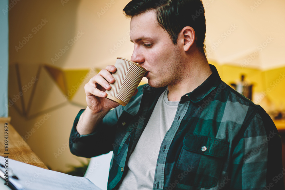 Poster clever hipster man drinking coffee while reading documents in folder for designing project, thoughtf