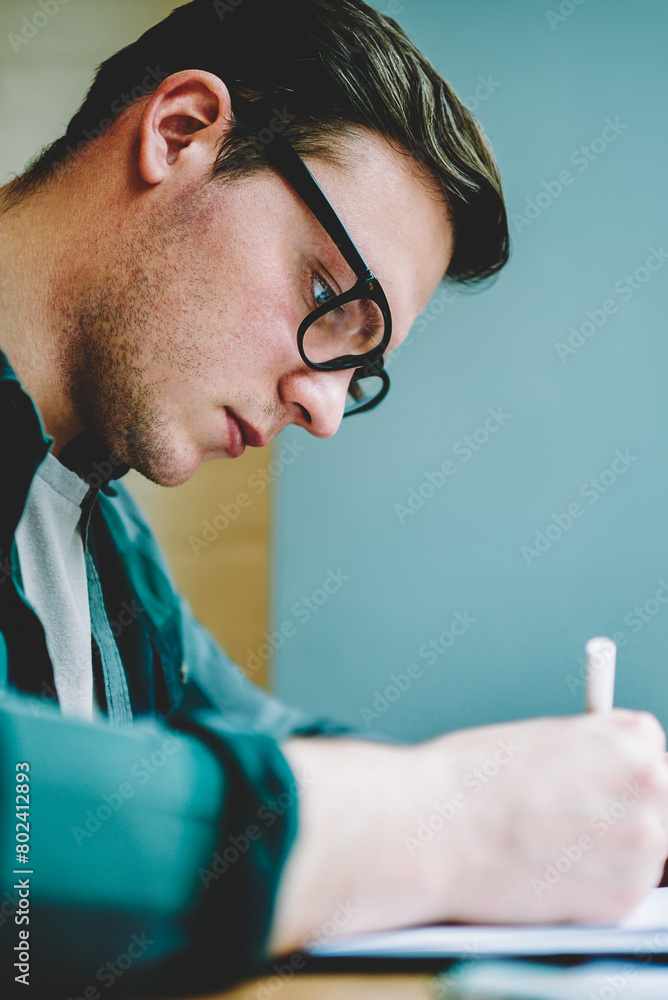 Poster close up view of smart student in spectacles writing essay for homework task concentrated on learnin