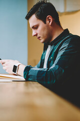 Pensive male student holding telephone connecting to wireless internet in coworking space for...