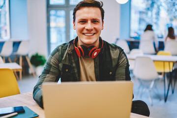 Portrait of cheerful hipster guy with headphones enjoying studying online language course on leisure using netbook in coworking space, prosperous male freelancer looking at camera during remote work