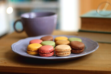 Purple plate filled with pastel macarons, cup of tea or coffee, vintage books and reading glasses on the table. Colorful bookcase in the background. Selective focus.