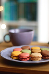 Purple plate filled with pastel macarons, cup of tea or coffee, vintage books and reading glasses on the table. Colorful bookcase in the background. Selective focus.