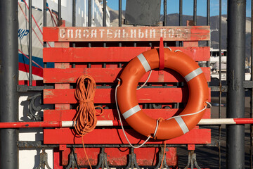 Orange lifebuoy with ropes hanging on a shield with a gaff. Inscription Rescue post
