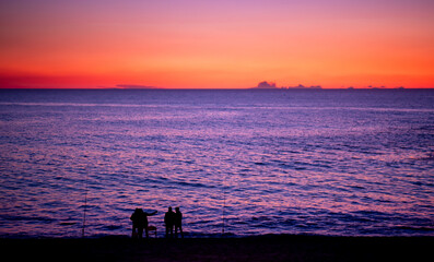 Group of men in silhouette fishing on beach at dusk, with red gradient sky