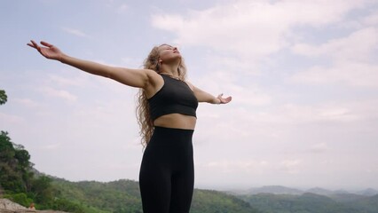 Woman black activewear stretches arms, breathes fresh mountain air. Female enjoys serene nature, finds peace hill. Curly-haired hiker relaxes, wilderness. Outdoor freedom, mindful breathing exercise