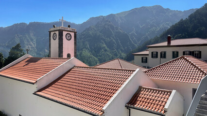 A church with red-tiled roofs and a clock tower is set against a backdrop of lush green mountains under a clear blue sky.