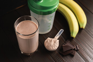 Protein milkshake cocktail in a glass, plastic measuring spoon with whey protein powder, shaker for prepare blended protein drink, chocolate cubes and banana fruit on a dark wooden background