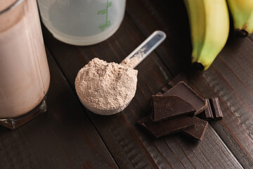Plastic measuring spoon with whey protein powder, milkshake cocktail in a glass, shaker for prepare blended protein drink, chocolate cubes and banana fruit on a dark wooden background