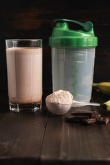 Plastic measuring spoon with whey protein powder, milkshake cocktail in a glass, shaker for prepare blended protein drink, chocolate cubes and banana fruit on a dark wooden background