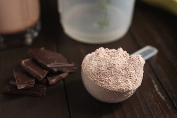 Plastic measuring spoon with whey protein powder, chocolate cubes, shaker for prepare milkshake cocktail or protein drink on a dark wooden background