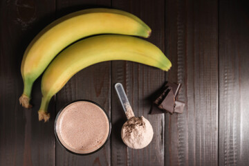 Chocolate milkshake cocktail in a glass, blended protein drink, plastic measuring spoon with whey protein powder and banana fruit on a dark wooden background, top view