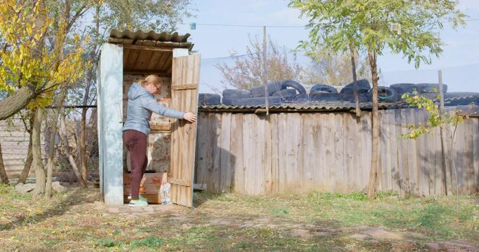 A woman is seen heading towards a traditional blue outhouse in a rustic setting, illustrating a natural and straightforward aspect of rural life