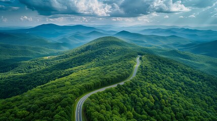 Scenic landscape view of a mountainous region covered in lush green forests. A winding road snakes through the dense tree-covered hills, leading towards the distant misty mountains in the background. 