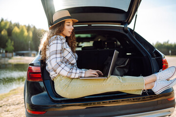 Travel and technology. Happy young woman working on laptop while sitting in trunk of car.