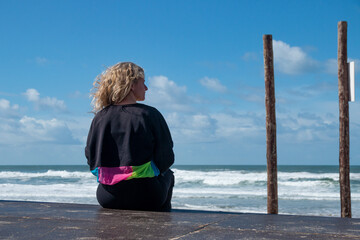 Blonde woman sitting on her back in front of the sea using her smartphone while the wind caresses...