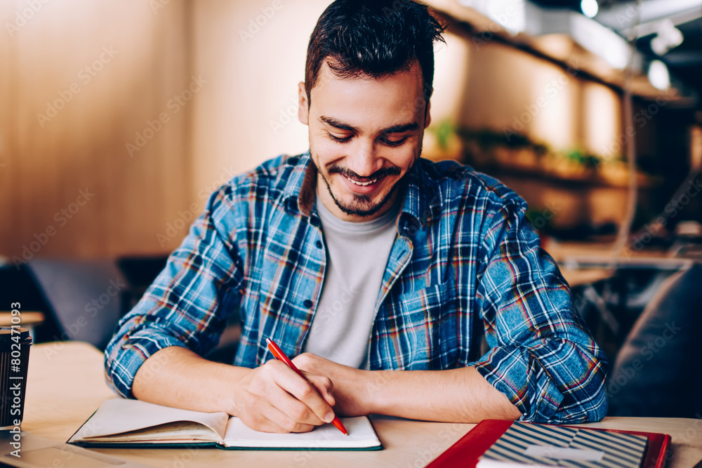 Poster smiling male blogger satisfied with having great idea for publication making notes sitting at deskto