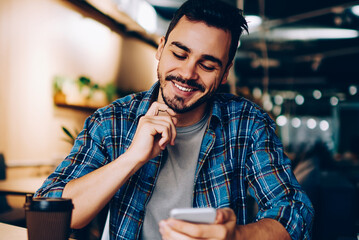 Smiling hipster guy reading new message in social networks confirming new friend on smartphone,...