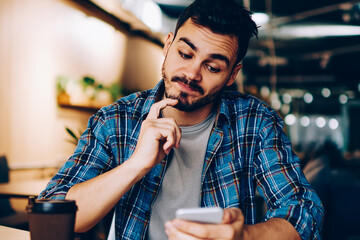 Contemplative male analyzing information about booking on website using cellular in coworking...