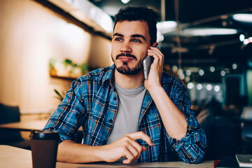Young hipster guy calling to friend using cheap mobile phone tariffs while sitting on coffee break,...