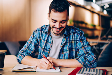 Smiling male blogger satisfied with having great idea for publication making notes sitting at desktop with netbook, positive student enjoying learning process doing homework task in college library