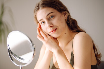 Beautiful woman looking in the mirror, touching her face with her hand, examines acne,...