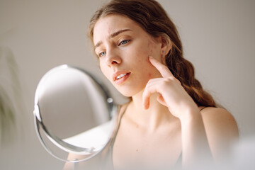 Beautiful woman looking in the mirror, touching her face with her hand, examines acne,...